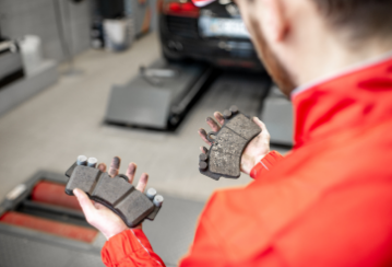 A mechanic holds a new brake pad in one hand and a worn one in the other while standing in a mechanic shop.